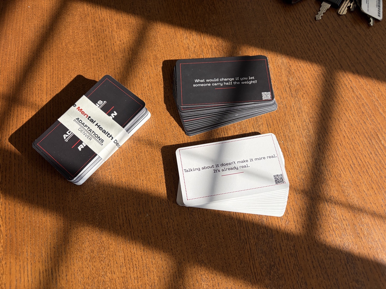 The MENtal Health Deck on a wooden table: the wrapped deck, a stack of black Reflection cards and a stack of white Reminder cards